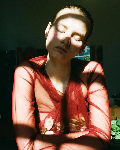 Young woman with closed eyes, bathed in light wearing a sheer red top with white flowers in an indoor setting. Contemplative portrait.