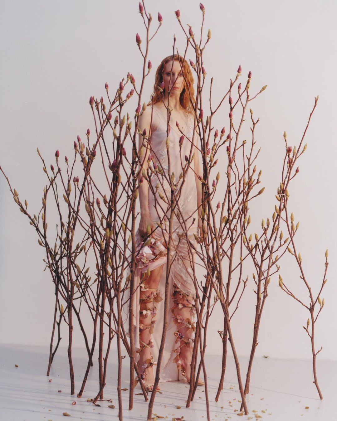 A person stands surrounded by budding branches wearing a draped, sleeveless gown with floral appliqués against a white background.