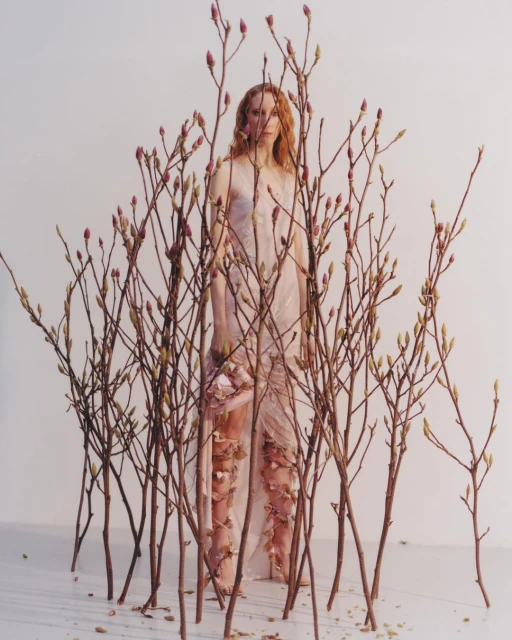 A person stands surrounded by budding branches wearing a draped, sleeveless gown with floral appliqués against a white background.
