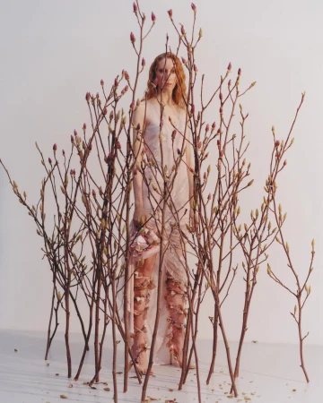 A person stands surrounded by budding branches wearing a draped, sleeveless gown with floral appliqués against a white background.