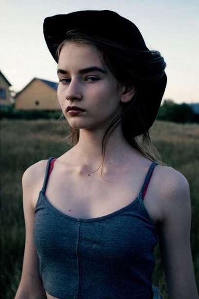 Portrait of a young woman with fair skin wearing a black hat, gray top, and red straps, standing outdoors in a grassy field at dusk.