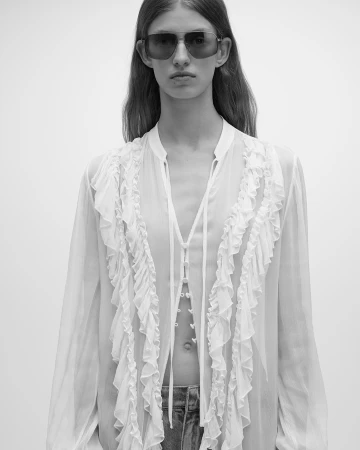 Model wearing dark sunglasses and a sheer white ruffled blouse against a neutral background