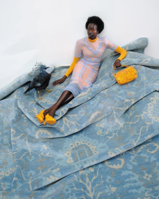 Dark-skinned woman poses on a patterned blue fabric with a black bird, wearing a yellow dress, shoes, and bag, on white background.