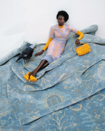 Dark-skinned woman poses on a patterned blue fabric with a black bird, wearing a yellow dress, shoes, and bag, on white background.
