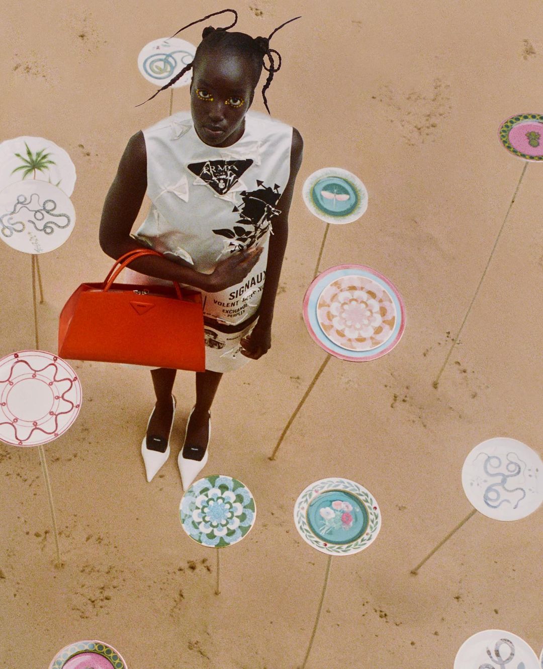 Woman in sleeveless top and skirt, holding a red bag, surrounded by decorative plates on sticks in a sand environment.