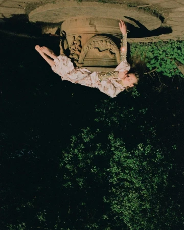 A young woman in a floral dress levitates upside down beneath an ornate stone pedestal, framed by lush greenery in a dark, mysterious scene.