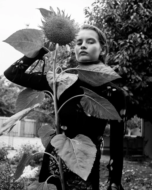 Black-and-white portrait of a young woman posing with a large sunflower outdoors in a garden setting, captured in natural light.