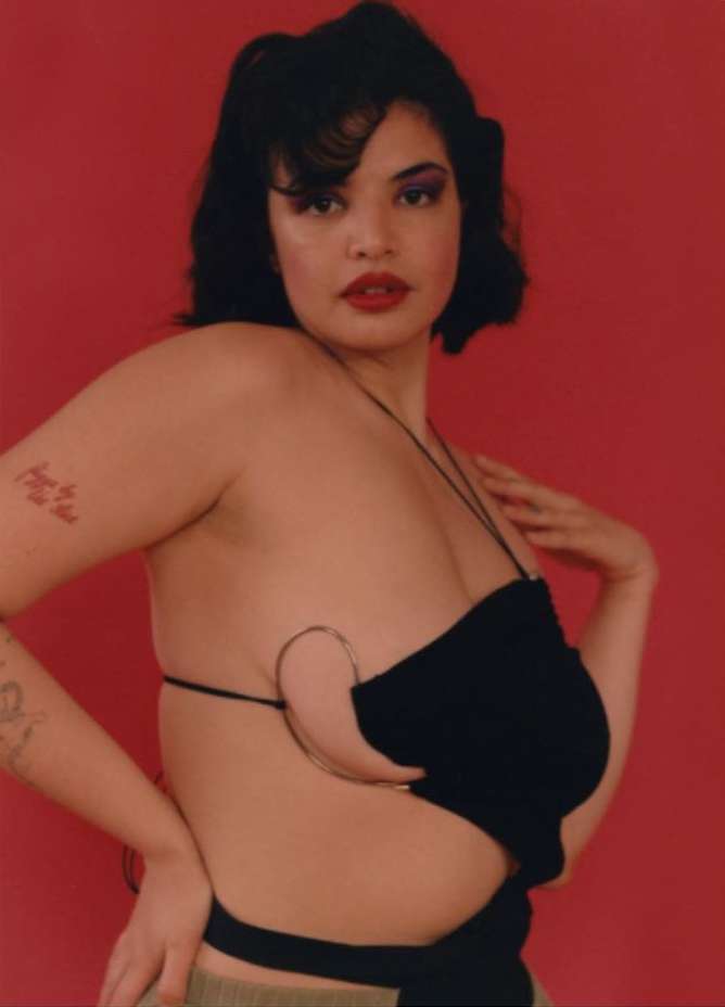 Portrait of a woman with dark hair and red lipstick, posing in a black top against a red background, showing shoulders and tattoos.