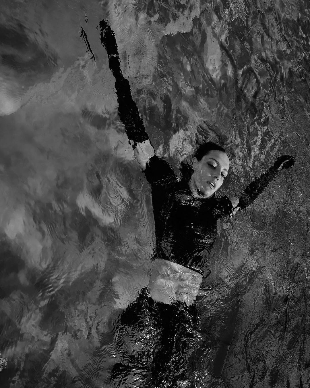 Black and white photo of a woman floating on her back in rippling water with her arms extended above her head.