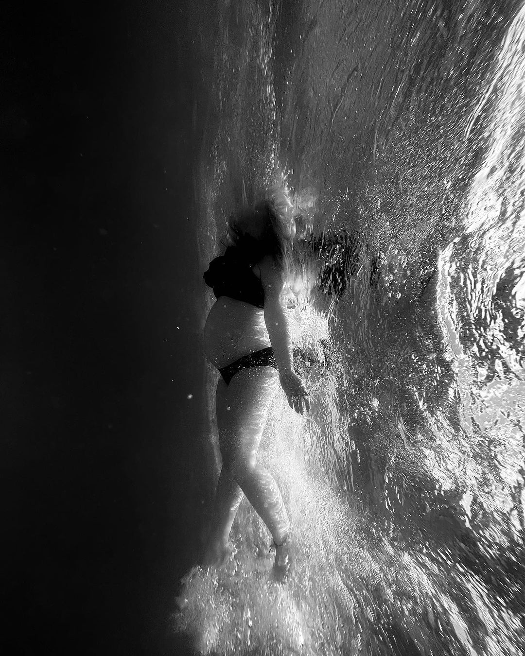 Pregnant woman underwater in black bikini swims in pool, captured in black and white photography, showcasing a peaceful water scene.