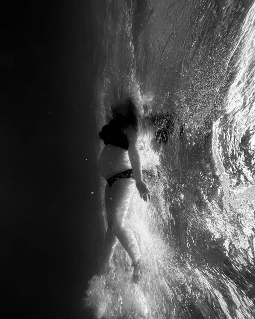 Pregnant woman underwater in black bikini swims in pool, captured in black and white photography, showcasing a peaceful water scene.