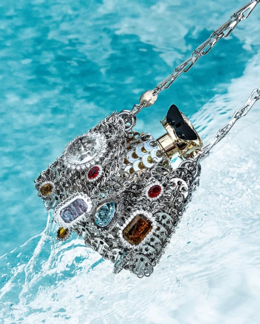 Floating silver handbag with gemstones and a perfume bottle inside, surrounded by light blue water with gentle ripples. Fashion accessory.