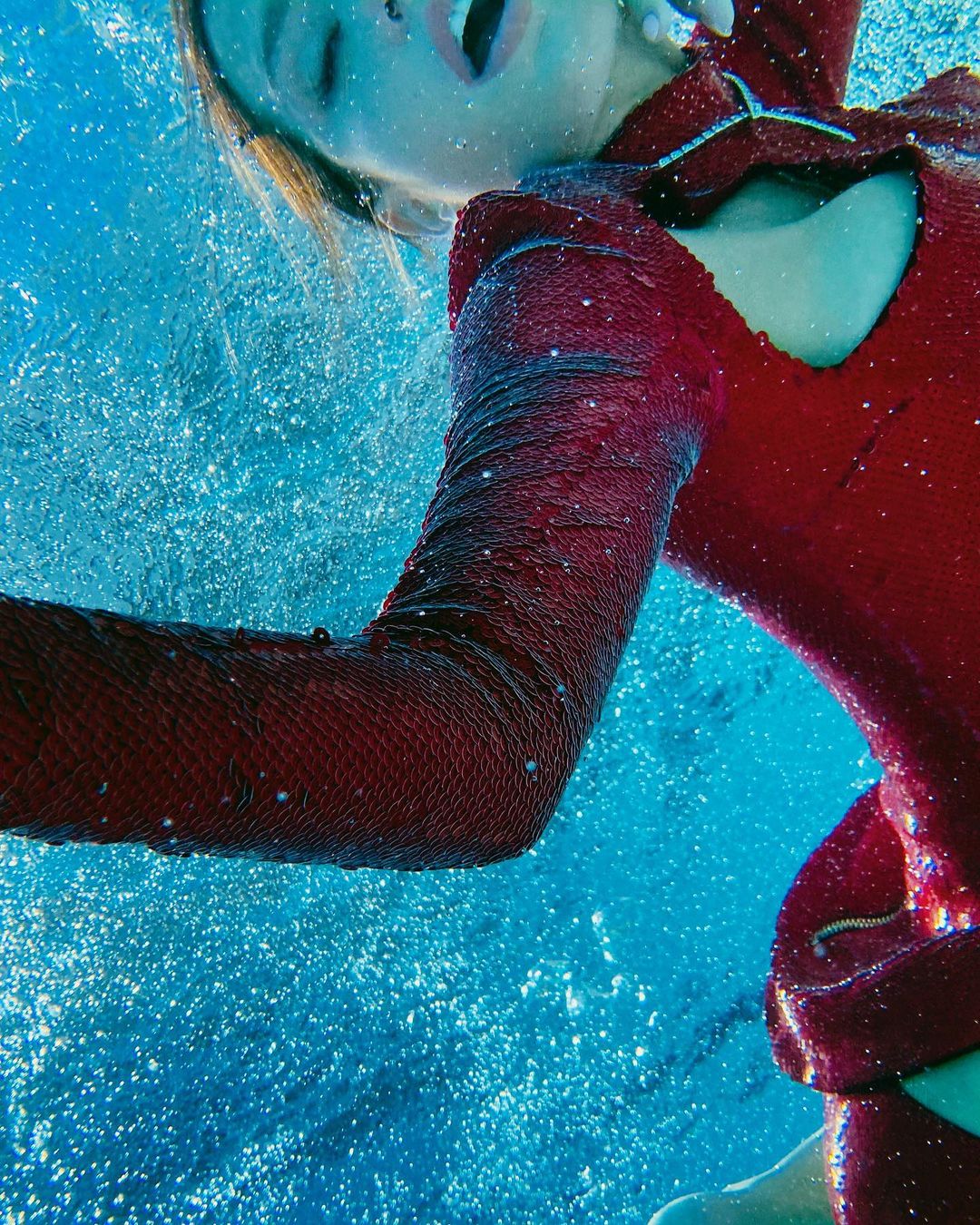 Underwater view of a person in a red sequined outfit with their mouth open amidst bubbles and blue water creating a dynamic image.