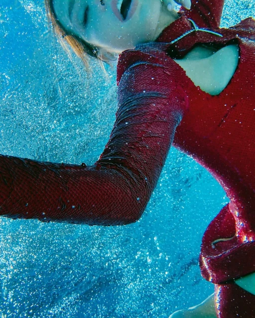 Underwater view of a person in a red sequined outfit with their mouth open amidst bubbles and blue water creating a dynamic image.