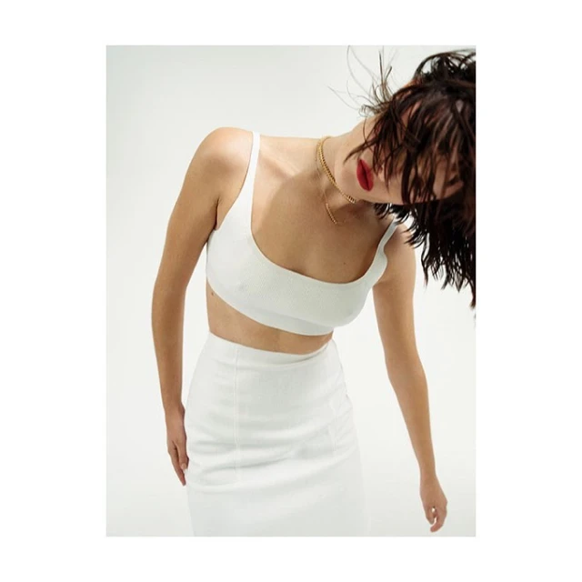 Brunette woman in a studio wearing a white two-piece outfit, gold chain, and red lipstick. Hair is windswept. White background.