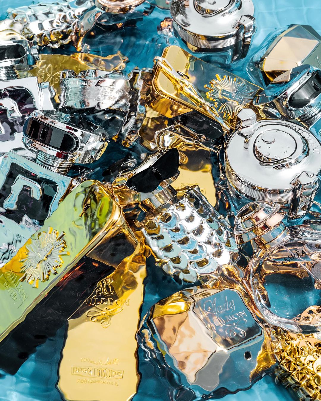 Luxury perfume bottles in gold and silver tones submerged in clear blue water creating a shimmering, high-end beauty product display.