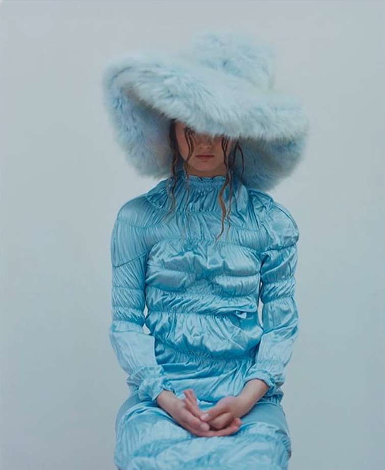 Woman in a blue dress and furry hat poses in a studio. The hat covers most of her face as she sits with hands folded.