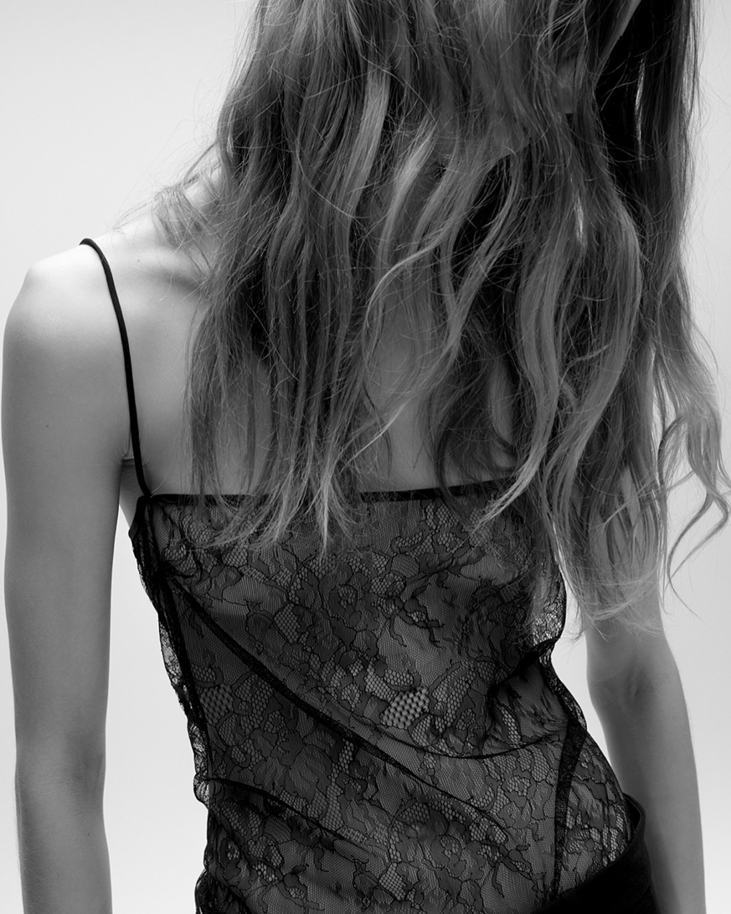 Back view of a slender adult female with flowing wavy hair, wearing a black lace top in a studio setting with a white background.