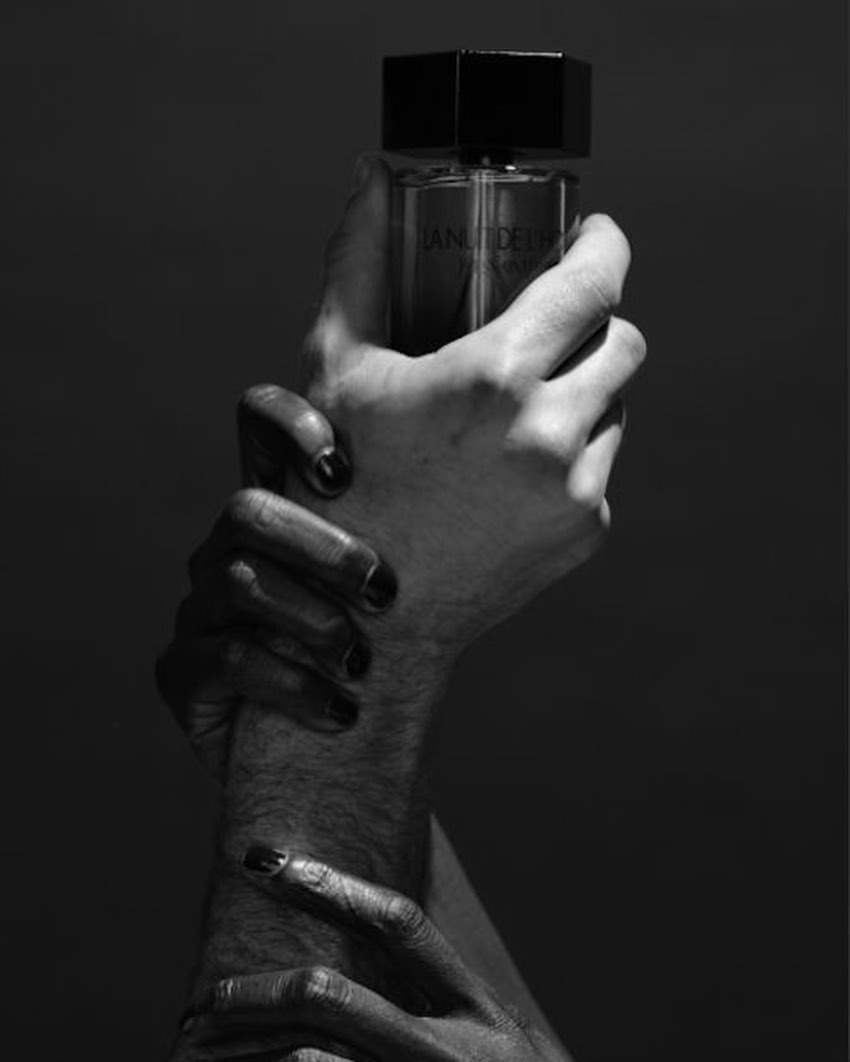 Black and white studio shot of a perfume bottle held by two hands with painted nails against a dark grey background.
