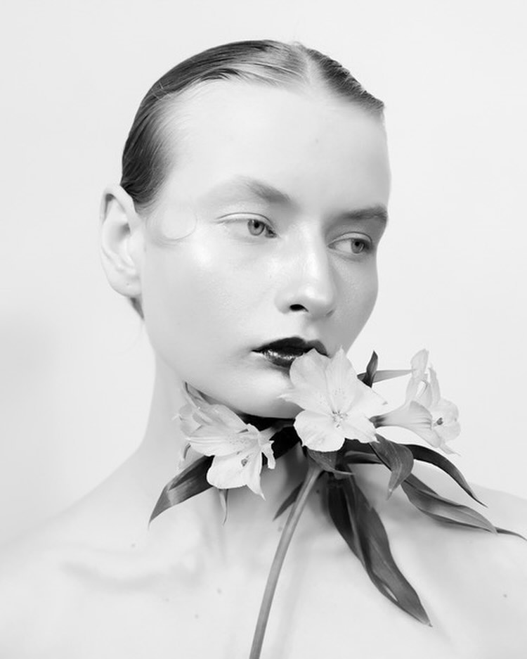 Black and white portrait of a female model with slicked-back hair and dark lipstick, holding white flowers to her neck against a white background.