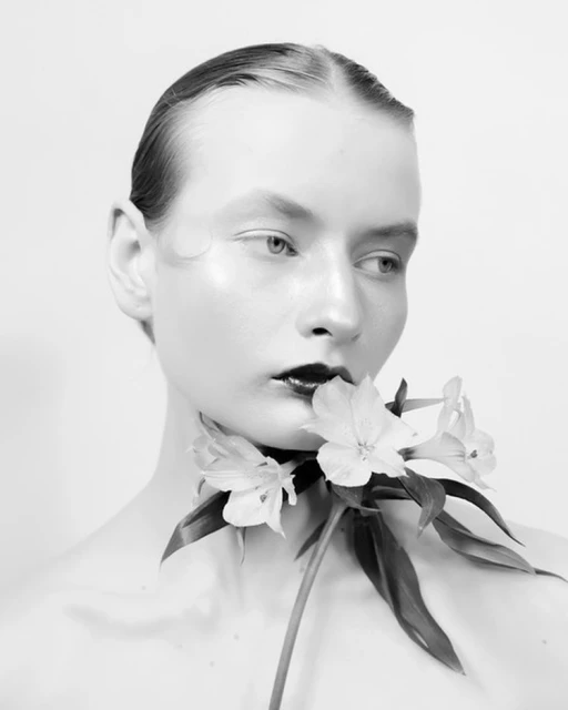 Black and white portrait of a female model with slicked-back hair and dark lipstick, holding white flowers to her neck against a white background.