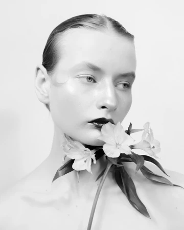 Black and white portrait of a female model with slicked-back hair and dark lipstick, holding white flowers to her neck against a white background.