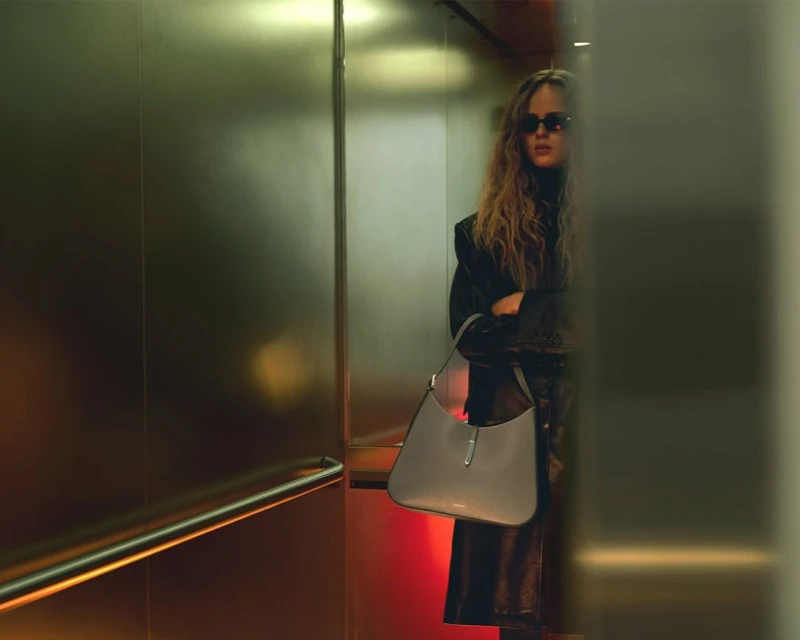 Woman with sunglasses stands in elevator holding purse, wearing a dark coat and turtleneck sweater.
