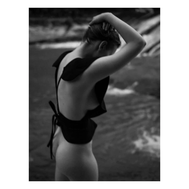 Monochrome portrait of a nude woman with dark fabric garment posing outdoors near a flowing river with soft, diffused lighting.
