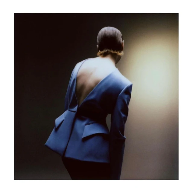 An adult woman with an open-back blue blazer poses in a studio with a soft light creating a dramatic effect and showcasing the garment's design.