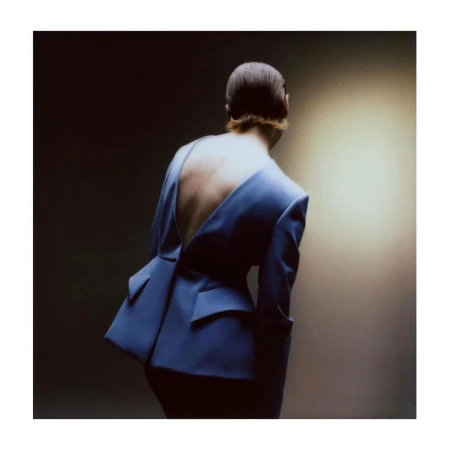 An adult woman with an open-back blue blazer poses in a studio with a soft light creating a dramatic effect and showcasing the garment's design.