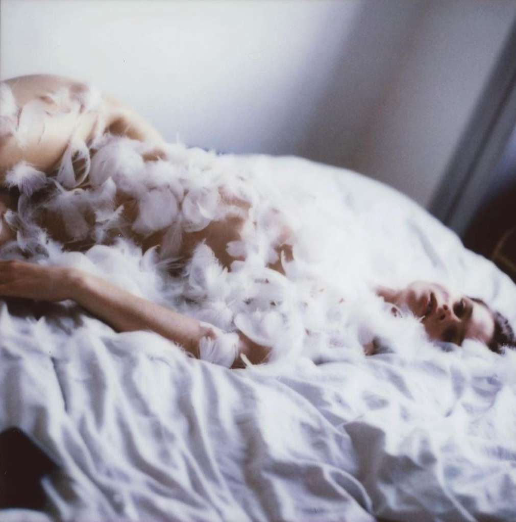 A woman in a feather-embellished garment is lying on a bed with white sheets, resting her head against a white headboard.