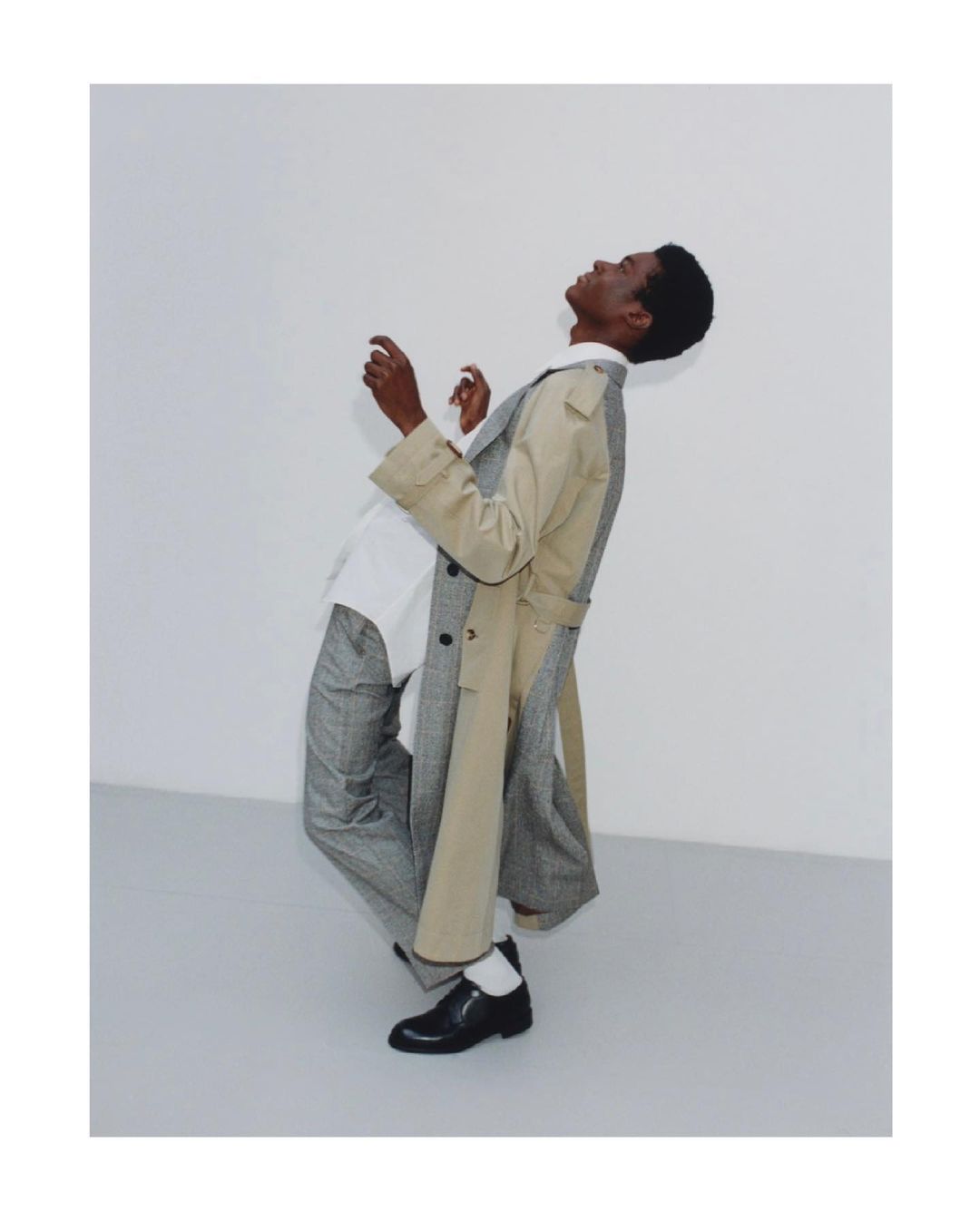 Young Black male model wearing a trench coat in a modern fashion pose against a white wall, shot in a photo studio with natural light.
