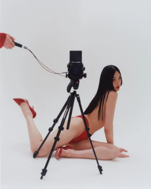 Asian woman posing on floor next to medium format camera in a photo studio wearing red heels and red bikini bottoms with white background.