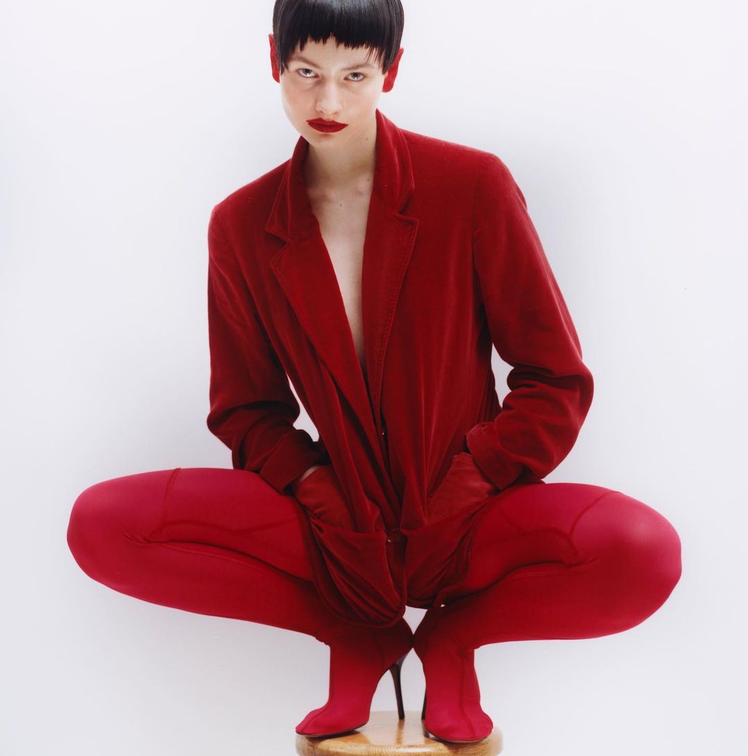 An adult female with a dark bowl cut wears a red velvet suit with matching red tights and heels, posed in a crouch on a wooden stool in a studio.