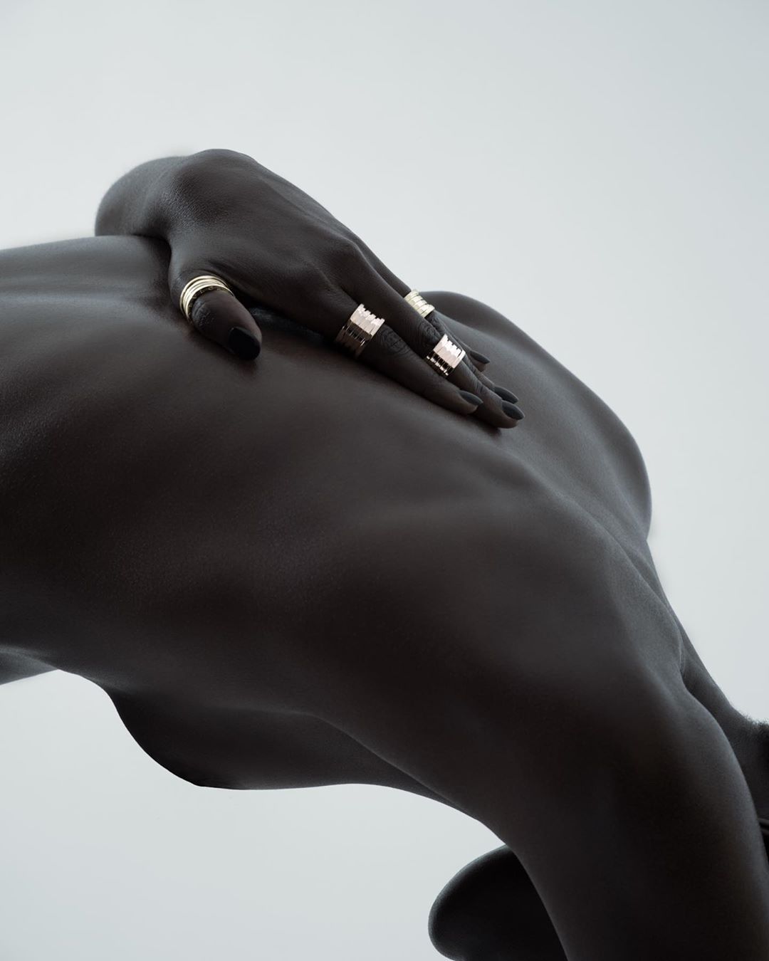 Dark-skinned woman’s back and hand with gold rings against a light background. Depicts jewelry, skin texture, and body contouring.