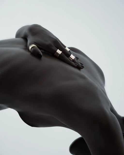 Dark-skinned woman’s back and hand with gold rings against a light background. Depicts jewelry, skin texture, and body contouring.