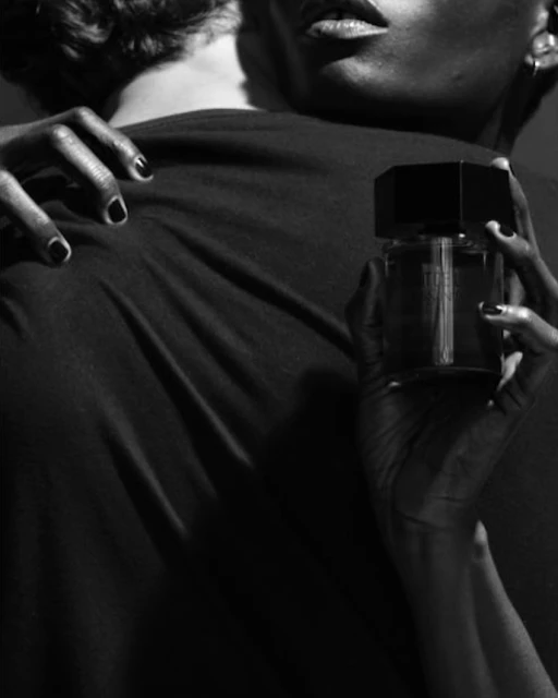 Close-up studio shot of a dark-skinned person holding a square glass perfume bottle, with another person in the background.