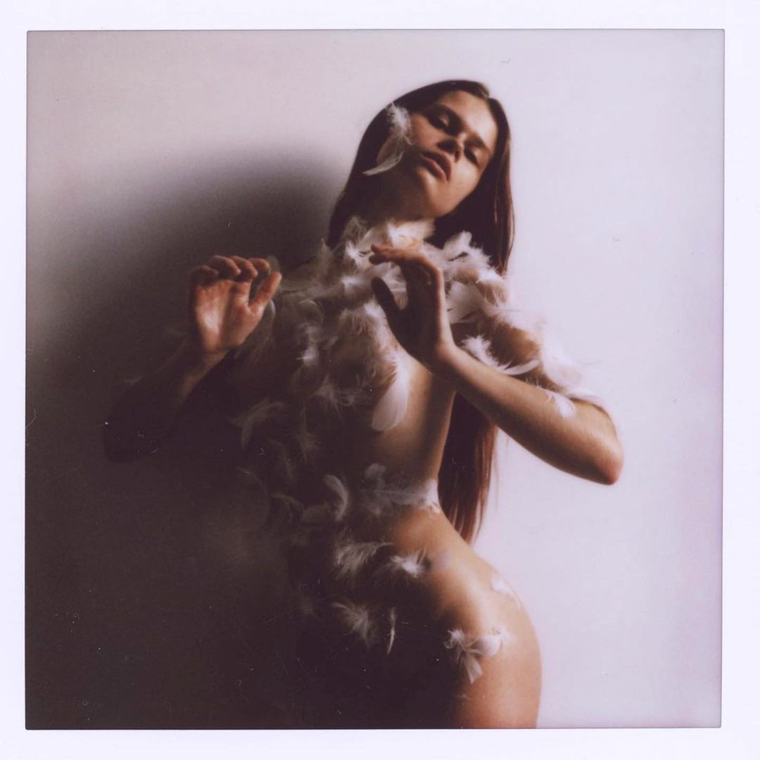Artistic nude portrait of a young woman adorned with white feathers, posing against a muted wall in soft, diffused lighting.