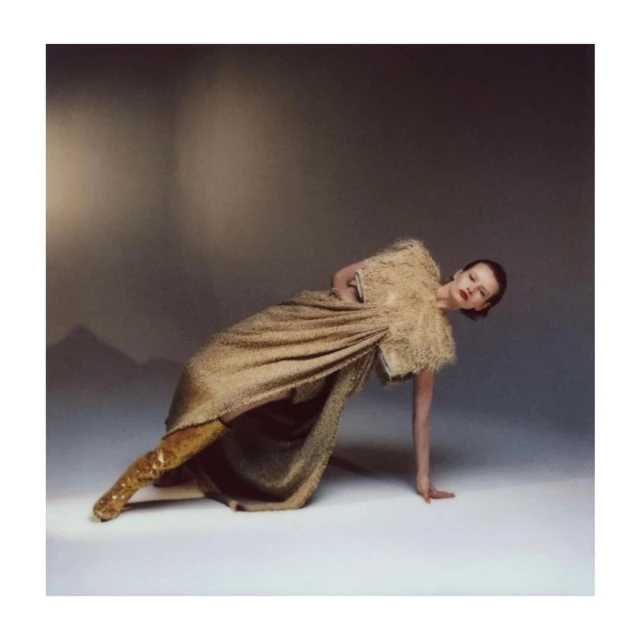 A single female model strikes a pose with one arm raised, wearing a gold dress with boots in a neutral studio environment.