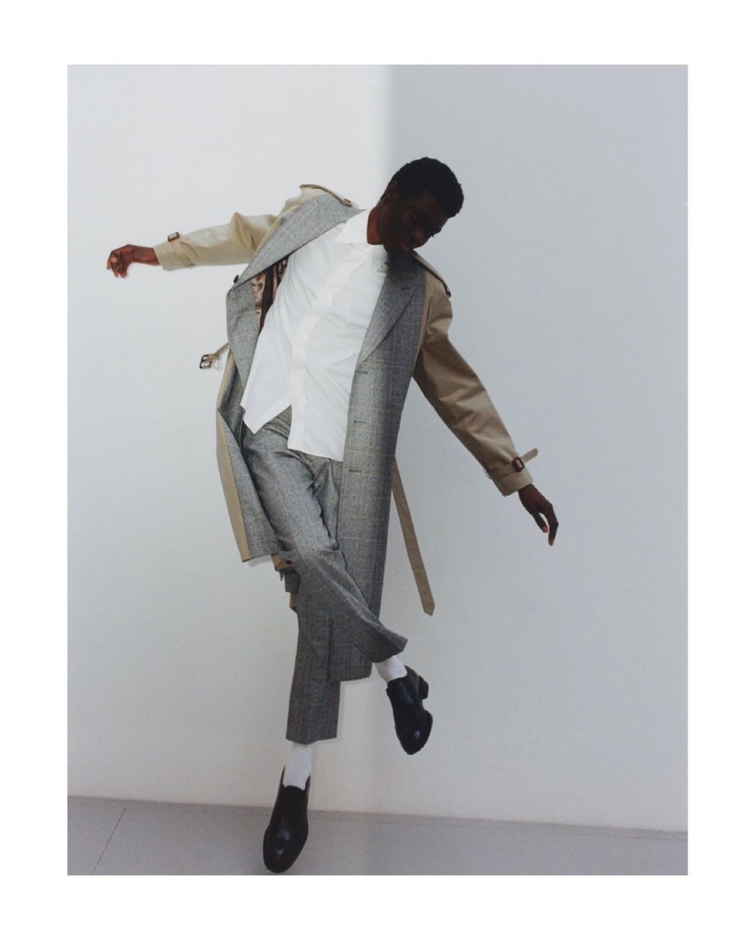A black male fashion model in mid-air wearing a beige trench coat, white dress shirt, grey patterned trousers, white socks and black loafers.