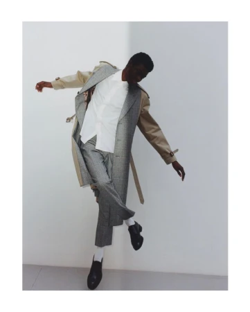 A black male fashion model in mid-air wearing a beige trench coat, white dress shirt, grey patterned trousers, white socks and black loafers.