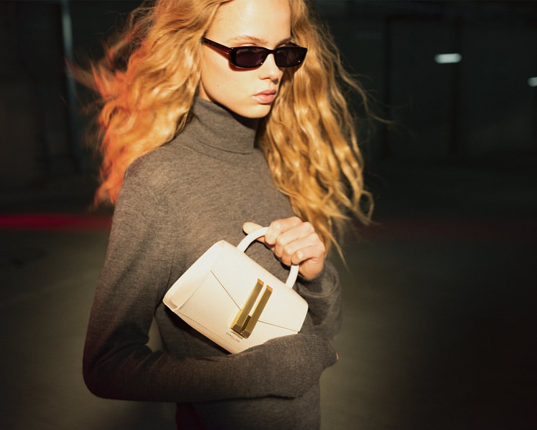A stylish young woman with wavy blonde hair, sunglasses, and a grey turtleneck sweater holds a modern white handbag in an indoor setting.