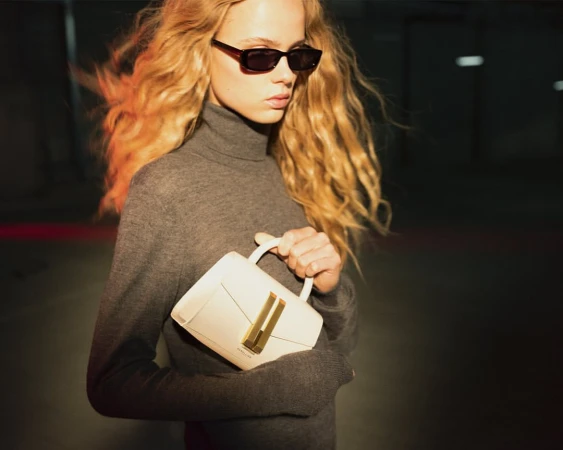 A stylish young woman with wavy blonde hair, sunglasses, and a grey turtleneck sweater holds a modern white handbag in an indoor setting.