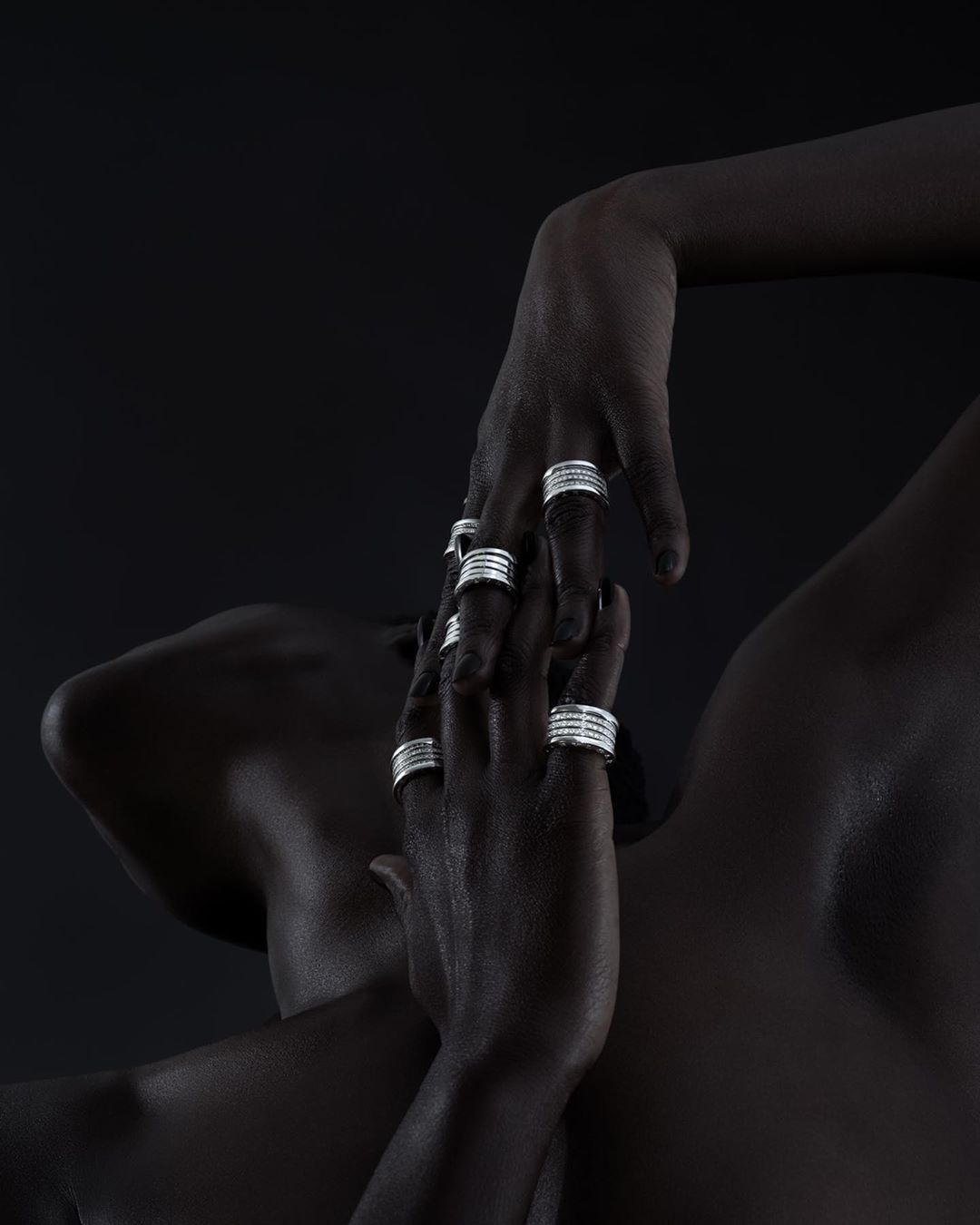 Close-up of a dark-skinned person's upper torso and hands adorned with silver rings against a dark backdrop, fashion beauty shot.