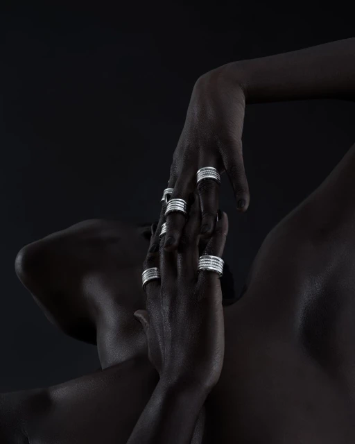 Close-up of a dark-skinned person's upper torso and hands adorned with silver rings against a dark backdrop, fashion beauty shot.
