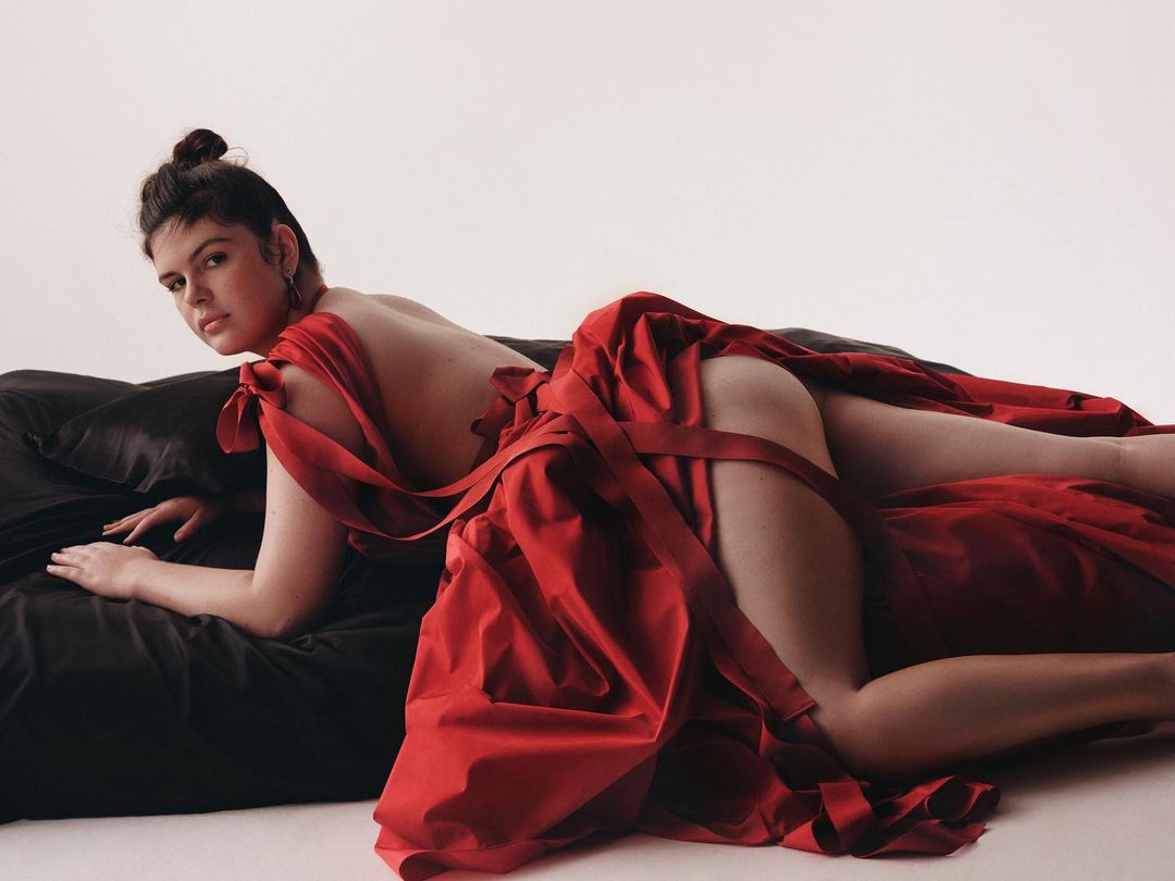Woman lying on a black satin surface in a red satin dress against a white background looking at the camera in a studio.