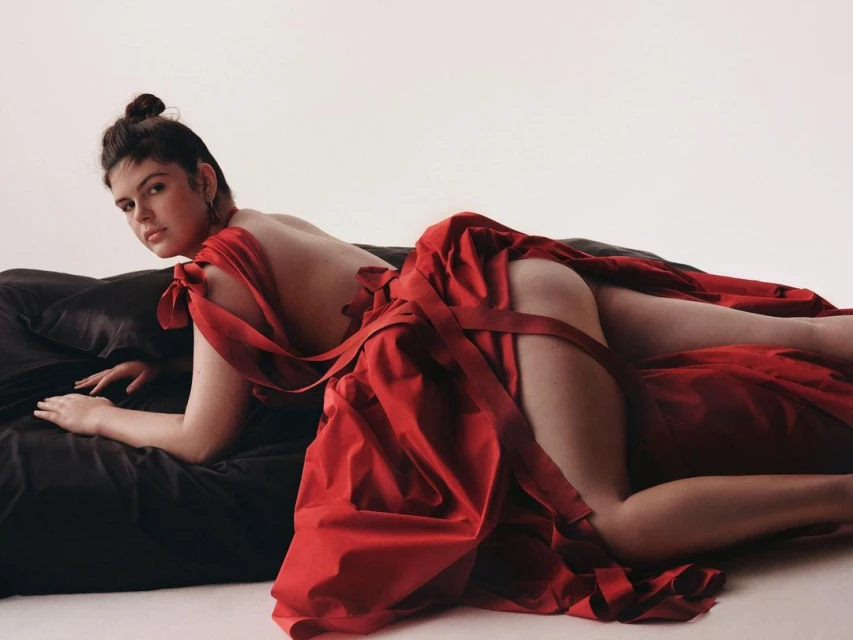 Woman lying on a black satin surface in a red satin dress against a white background looking at the camera in a studio.
