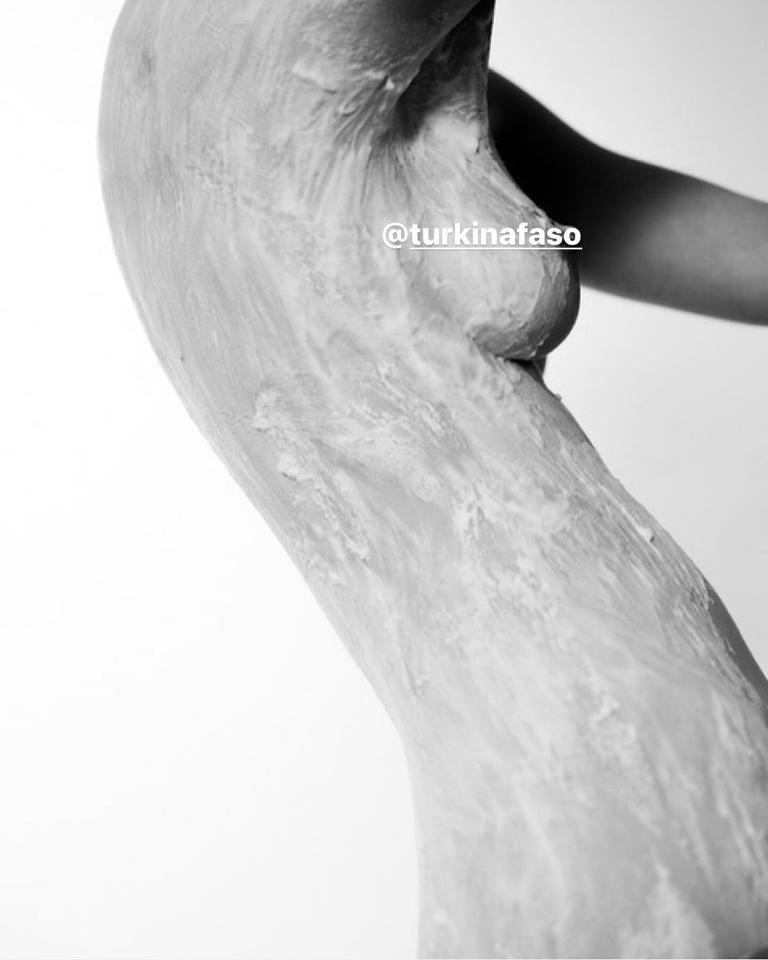 Black and white image of an adult woman's upper torso and arm, textured with what appears to be clay or paint, against a white backdrop.