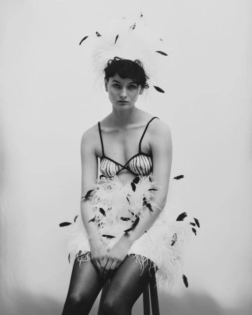 Monochrome portrait of a young woman in a feathered outfit, seated on a stool, looking at the camera against a plain background.