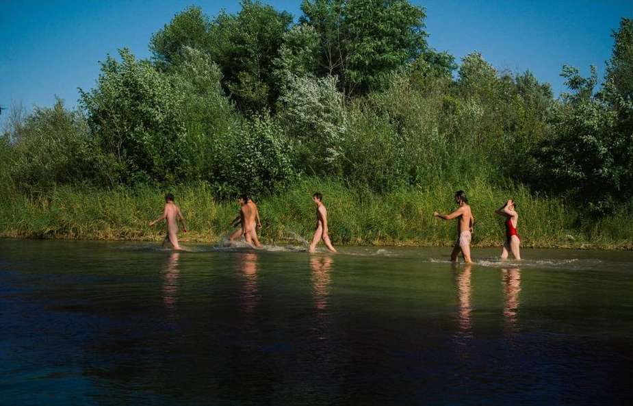 Five people wading in a river bordered by lush green vegetation and trees beneath a clear blue sky, enjoying nature on a sunny day.